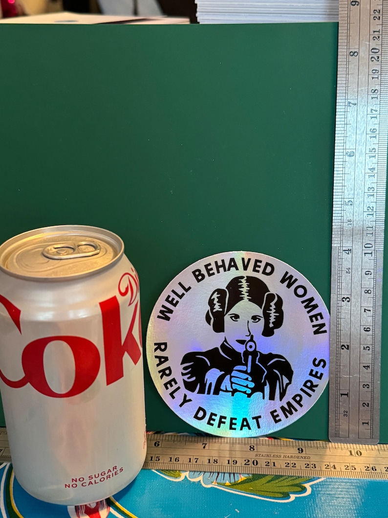 May include: A silver holographic sticker with a black and white image of a woman holding a gun. The sticker reads "Well Behaved Women Rarely Defeat Empires".