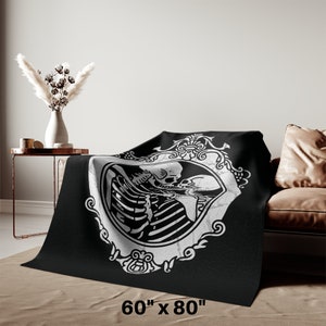 May include: Black and white throw blanket featuring a skeleton couple kissing inside an ornate frame. The blanket is 60 inches by 80 inches.