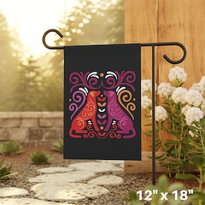 May include: A black garden flag featuring a colourful design of two dogs facing each other. The dogs are outlined in red, orange, and purple. The design is surrounded by white and black floral patterns. The flag measures 12 inches by 18 inches.
