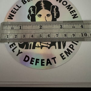 May include: A silver holographic sticker with black text that reads "Well Behaved Women Rarely Defeat The Empire". The sticker features a black and white illustration of Princess Leia from Star Wars.