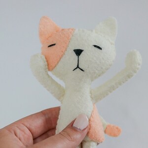 May include: A white felt cat with peach-colored ears and a peach-colored patch on its belly. The cat has black eyes and a black nose.