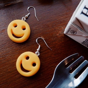 May include: A pair of yellow smiley face earrings with silver hooks. The earrings are round with two eye holes and a curved mouth. The earrings are on a dark wooden surface with a fork and a package of food.