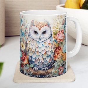 May include: White ceramic mug with a colorful owl illustration. The owl is surrounded by flowers and foliage in shades of pink, orange, yellow, and blue. The mug has a white handle.
