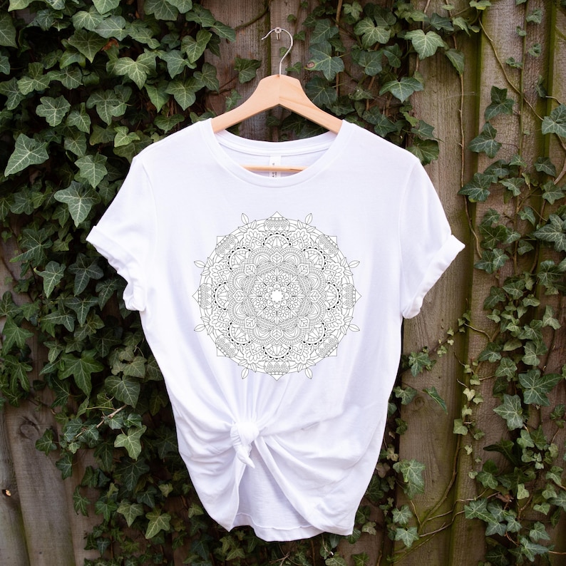 Puede incluir: Una camiseta blanca con un dise&ntilde;o de mandala en blanco y negro detallado en la parte delantera. La camiseta est&aacute; colgada en una percha de madera sobre un fondo de follaje verde. La camisa est&aacute; anudada a la cintura.