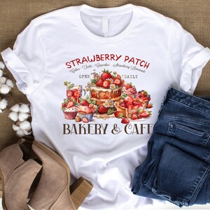 Pode incluir: Camiseta branca com um design gráfico de uma padaria chamada "Strawberry Patch Bakery & Cafe". O design inclui um bolo, cupcakes e morangos. O texto "Aberto diariamente" também está incluído no design.