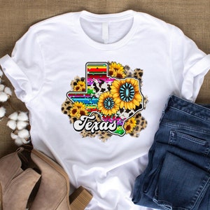May include: White t-shirt with a colorful graphic of the state of Texas. The graphic features a rainbow striped outline of the state, a cow print, and sunflowers with turquoise accents. The word "Texas" is written in a retro font below the graphic.