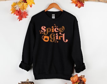 May include: Black crewneck sweatshirt with the text "Spice Girl" in retro-style lettering. The design includes a coffee mug with a pumpkin graphic and star accents. The sweatshirt is hanging on a wooden hanger.