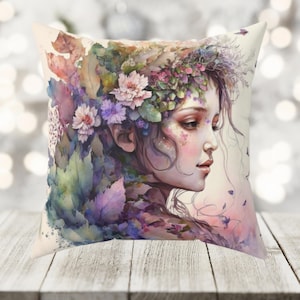 Puede incluir: Una almohada decorativa con una pintura de acuarela de la cara de una mujer rodeada de flores y hojas. La mujer tiene el pelo largo y oscuro y lleva una corona de flores. El fondo es de un blanco suave.