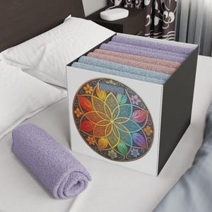 May include: A white storage box with a colorful floral mandala design on the front. The box is filled with folded towels in shades of purple, pink, and blue. A rolled-up purple towel rests on the bed.