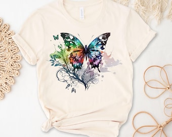 Peut inclure: Un t-shirt crème avec un motif de papillon coloré. Le papillon a des ailes bleues, vertes, rouges, oranges et violettes. Le papillon est entouré de motifs floraux noirs.