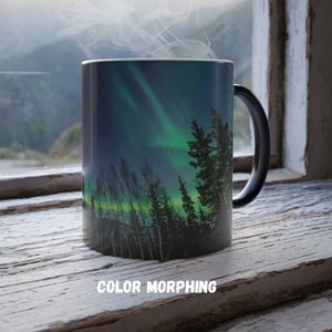 May include: A color-morphing mug featuring a vibrant image of the Northern Lights. The design showcases a green and blue aurora borealis over a silhouetted forest. The mug has a black handle and the text "COLOR MORPHING" at the bottom.