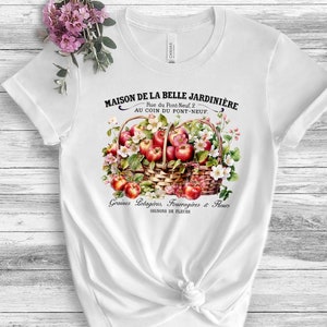 May include: White t-shirt with a vintage-style illustration of a basket overflowing with red apples and white flowers. The text "MAISON DE LA BELLE JARDINIERE" is printed above the basket, and "Graines Rotagères, Fourragires & Fleurs" is printed below the basket.