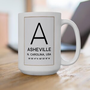 May include: White ceramic mug featuring a large black letter "A" and the city name "ASHEVILLE" in black text. Below the city name is "N. CAROLINA, USA" and coordinates. The mug is on a wooden surface.