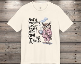 Puede incluir: Una camiseta blanca con un búho de dibujos animados con pijama y sosteniendo una taza de café. El texto en la camiseta dice "Not a morning bird, not even a night owl... just tired."