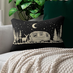 May include: Black rectangular throw pillow with a cream-colored illustration of a camper, deer, and trees under a starry night sky with a crescent moon. The pillow is on a light gray couch with a cream-colored blanket.