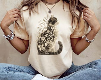 Könnte beinhalten: Cremefarbenes T-Shirt mit einer detaillierten Illustration einer Katze, die mit floralen Mustern verziert ist. Die Katze ist in Grau- und Weißtönen gehalten, mit aufwendigen floralen Designs. Das Shirt ist ein Freizeitartikel.