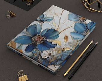May include: A hardcover journal with a floral design featuring large blue flowers with brown centers and gold accents. The journal is open, revealing blank pages. Gold and black pencils and gold binder clips are nearby.