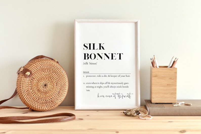 Puede incluir: Una impresi&oacute;n enmarcada con la palabra "SILK BONNET" en negrita en negro, seguida de la pronunciaci&oacute;n "/silk b&auml;nət/" y la definici&oacute;n: "sustantivo 1. protector, amigo fiel y guardi&aacute;n de tu cabello. 11. incluso cuando se desliza y desaparece misteriosamente por la noche, siempre te quedar&aacute;s a su lado. cuidado del cabello de primera"