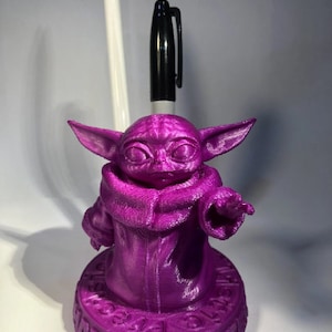 May include: A purple 3D printed pen holder shaped like the character Baby Yoda from the Star Wars series. The holder has the text "Do what you see" on the base.