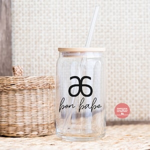 May include: A clear glass jar with a bamboo lid and a black and white logo that says "bon babe" and a symbol that looks like a double "a". The jar has a glass straw inside and is sitting on a woven surface.