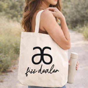 May include: A cream-coloured tote bag featuring a black logo and the words "fizz dealer" in a script font. A light-coloured water bottle is held in the hand. The bag has a simple design and is carried over the shoulder.