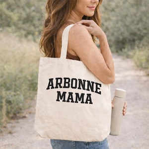 May include: A cream-coloured tote bag with the text "ARBONNE MAMA" in bold black letters. The bag has a shoulder strap and is being carried outdoors. A light grey water bottle is held in the same hand.