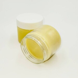 May include: Two jars of yellow balm, one with a white lid and one with a clear lid. The jars are on a white surface.