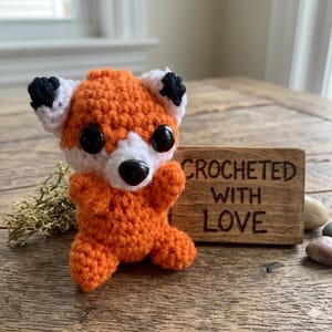 May include: A crocheted orange fox with white and black details. The fox has large black eyes and is positioned in front of a wooden sign that reads "CROCHETED WITH LOVE". A handmade item.