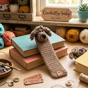May include: A light brown crocheted dog bookmark featuring a three-dimensional head with floppy ears and a long rectangular body. The bookmark includes a small leather tag at the bottom and is displayed resting on two stacked hardcover books.