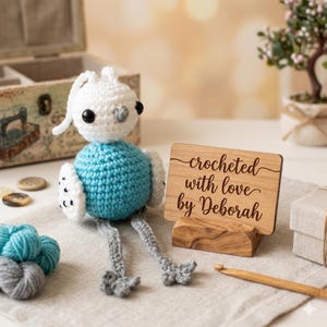 May include: A handcrafted crochet bird with a white head, turquoise body, and grey legs. It rests on a neutral surface beside a wooden sign reading "crocheted with love by Deborah."