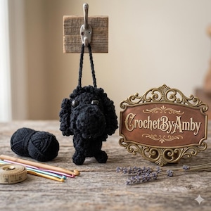 May include: A black crocheted dog ornament suspended from a hook. The dog features a textured design and button eyes. A ball of black yarn, crochet hooks, and a tape measure are on a wooden surface. A sign reading "CrochetByAmby" is also visible.