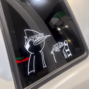 以下が含まれることがあります： サングラスをかけた漫画のキャラクターの白いビニールステッカーが車の窓に貼られています。1つは鳥のような姿、もう1つはアライグマのようなキャラクターです。ステッカーは、暗い窓に対して白で縁取られています。