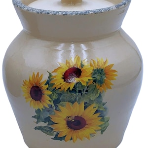 May include: A beige ceramic jar with a lid, adorned with a vibrant sunflower design. The jar has a rounded shape with a slightly flared rim and a small, round lid. The sunflowers are yellow with dark red centers and green leaves.