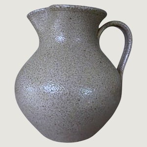 May include: A beige ceramic pitcher with a speckled, textured surface. The vessel features a rounded, bulbous body, a wide flared rim with a distinct pouring spout, and a single curved handle attached to the side of the neck and upper body.