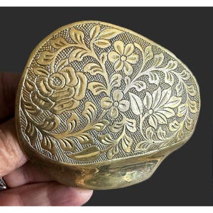 May include: An irregular, rounded brass object featuring an intricate, engraved floral pattern. The surface displays a large central rose, smaller blossoms, and detailed leaves set against a cross-hatched background, with a smooth, plain lower section.