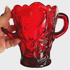 May include: A ruby red glass sugar bowl with two handles. The bowl features a heart and leaf pattern. The rim has a scalloped edge. The glass has a glossy finish. The sugar bowl is a vintage item.