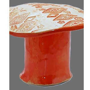May include: A ceramic mushroom-shaped decorative object. The stem is a glossy, vibrant orange, while the cap is white with a repeating pattern of orange diamond and floral designs. The object has a whimsical, artistic aesthetic.