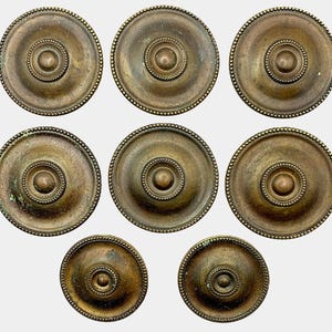 May include: A collection of eight antique bronze-colored cabinet knobs. Each knob features a central raised circle, surrounded by concentric rings and a beaded edge. The knobs vary slightly in patina, suggesting age and use. Ideal for vintage furniture restoration.