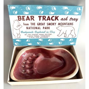 May include: A ceramic ash tray shaped like a bear footprint with a glossy maroon interior and a beige rim, presented in a box labeled BEAR TRACK ash tray from THE GREAT SMOKY MOUNTAINS NATIONAL PARK, Footprints Captured in Clay, PIGEON FORGE.