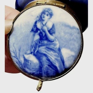 May include: A round metal compact case featuring a blue and white porcelain-style inlay on the lid. The inlay depicts a seated figure in a flowing garment against a soft, painterly background. The case has a metallic gold-toned rim and hinge.
