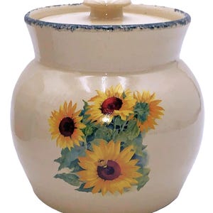 May include: A cream-colored ceramic jar with a lid and a knob. The jar is decorated with a bouquet of sunflowers in yellow, red, and green. The rim of the jar and lid have a blue trim. A decorative kitchen item.