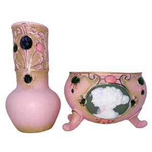 May include: A pink vase and a matching footed bowl featuring gold-toned accents, dark green and pink cabochon-style embellishments, and a central oval cameo relief depicting two white profiles against a dark green background on the bowl.