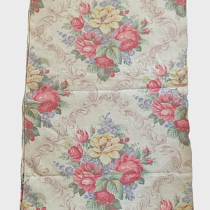 May include: Cream-colored fabric with a repeating floral pattern. Large bouquets of pink and yellow roses are interspersed with smaller blue flowers and green leaves. The background features a scrolling design.