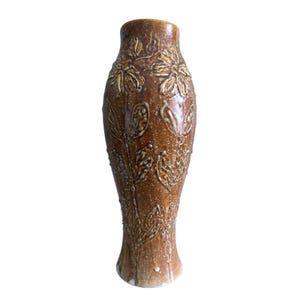 May include: A tall, cylindrical ceramic vase with a glossy, speckled brown glaze. The surface features a raised, embossed relief pattern of stylized flowers and foliage that wraps around the body, creating a textured, tactile appearance.