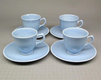 May include: Four light blue teacups and saucers are arranged on a neutral surface. Each cup has a rounded shape with a handle, and sits on a matching saucer. The set has a simple, classic design with a white rim.