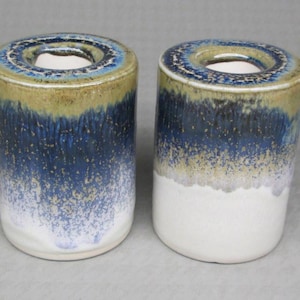 May include: Two ceramic candle holders with a cylindrical shape. The holders feature a color gradient, transitioning from a white base to a speckled blue and gold upper section. The top rim is a darker blue with a circular opening.