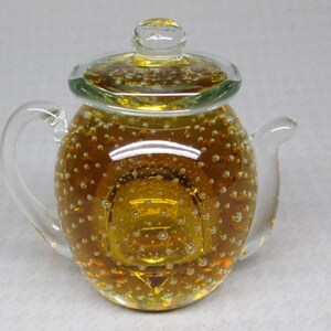 May include: A clear glass teapot with a rounded body, handle, and spout. The teapot is filled with a golden-yellow liquid and numerous tiny bubbles. The lid is also clear glass. The overall design is elegant and decorative.