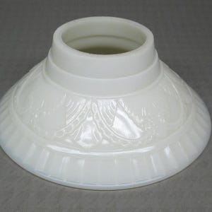 May include: A white, opaque glass lampshade with a flared, conical shape. The shade features a detailed, embossed floral pattern around the base and vertical ribbed detailing. The top has a cylindrical opening.