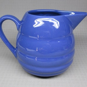 May include: A blue ceramic pitcher with a curved handle and a spout. The pitcher has a ribbed design around the body. The pitcher is a solid blue color and appears to be made of ceramic.