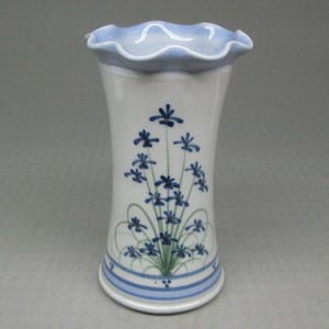 May include: A white ceramic vase with a flared shape and a ruffled, light blue rim. The vase features a hand-painted design of blue flowers and green stems. The base has a blue stripe. The vase is approximately 8 inches tall.